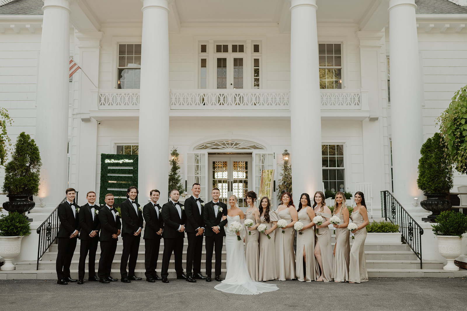 The full wedding party poses on the steps of a grand white mansion, showing elegance and joy.